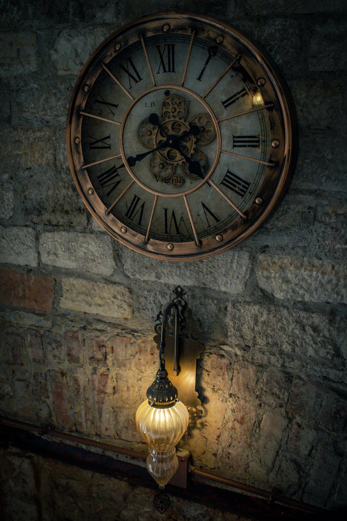 Vintage wall clock with Roman numerals and a glowing antique lamp on stone wall.
